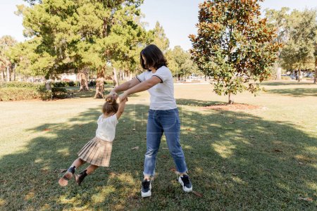 full length of brunette woman in jeans playing with toddler daughter in park of Miami 