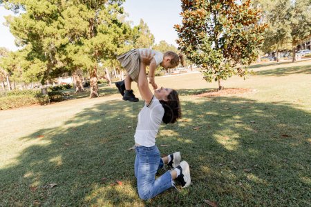 full length of cheerful mother in jeans lifting toddler daughter while playing in park of Miami 