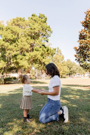 fröhliche Mutter auf Knien stehend und Händchen haltend mit ihrer kleinen Tochter beim Spielen im Park von Miami 