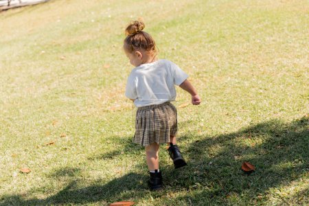 back view of toddler girl in white t-shirt and skirt walking in boots on green grass 