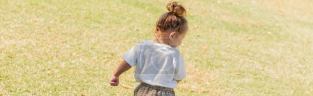 back view of toddler girl in white t-shirt walking on green grass, banner 
