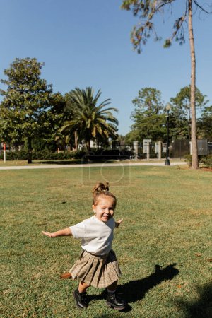 joyful toddler girl standing with outstretched hands while playing in green park 