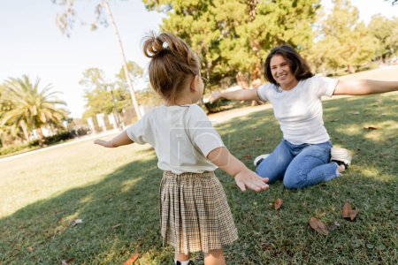 Kleinkind steht mit ausgestreckten Händen neben glücklicher Mutter beim gemeinsamen Spielen im Park von Miami 