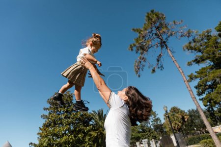 happy mother lifting toddler daughter against green trees and clear sky 