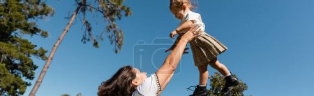 happy mother lifting toddler daughter against green trees and clear sky, banner 