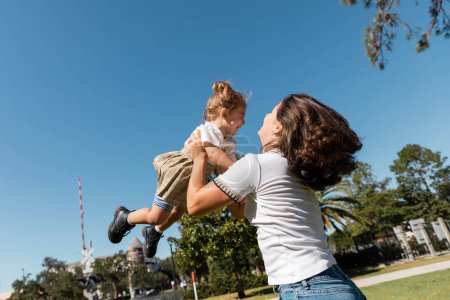 glückliche Mutter hebt Kleinkind-Tochter gegen grüne Bäume und wolkenlosen Himmel 