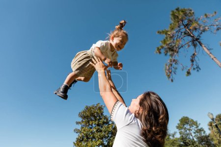 Téléchargez les photos : Mère soulevant heureux tout-petit fille contre les arbres verts et ciel sans nuages - en image libre de droit