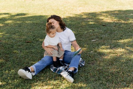 mother kissing baby girl while holding smartphone and sitting on lawn 