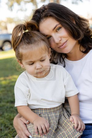 portrait of brunette mother looking at camera near toddler daughter 