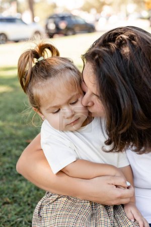portrait of brunette mother kissing cheek of daughter in white t-shirt 