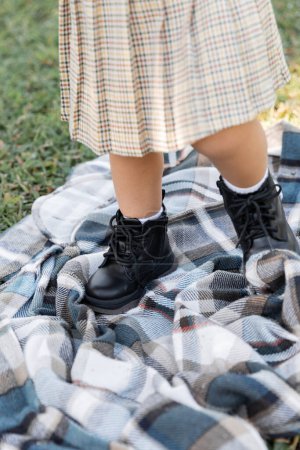 cropped view of baby girl in checkered skirt and boots standing on blanket near grass 
