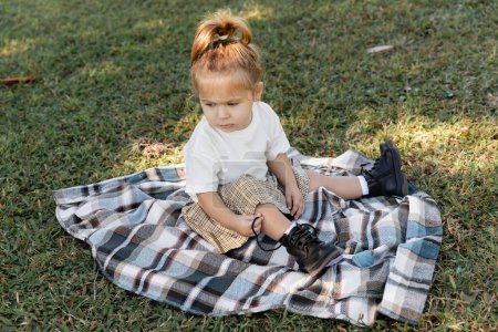 niña con falda a cuadros y botas sentadas en una manta durante el picnic 