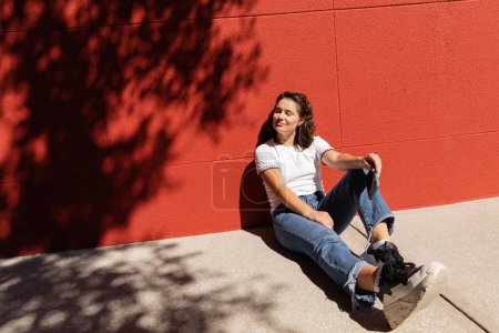 Téléchargez les photos : Femme gaie en t-shirt blanc et jeans bleu assis près du bâtiment avec mur rouge - en image libre de droit