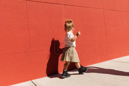 toddler girl in checkered skirt and white t-shirt walking near building with red wall 