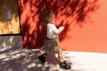 happy toddler girl in checkered skirt and white t-shirt walking near building with red wall 