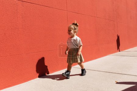 cheerful toddler girl in checkered skirt and white t-shirt walking near building with red wall 