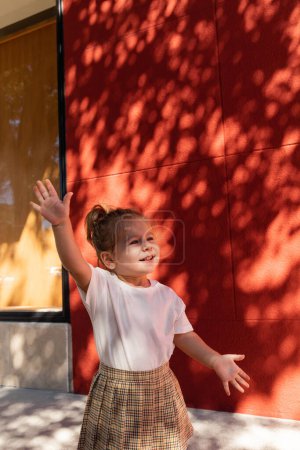 glückliches Mädchen in kariertem Rock und weißem T-Shirt, das in der Nähe eines Gebäudes mit roter Wand winkt 
