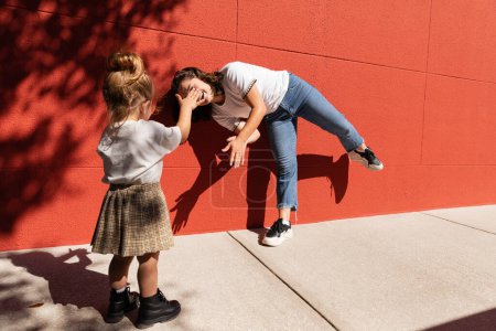 Kleinkind und glückliche Mutter spielen zusammen in der Nähe des roten Gebäudes 