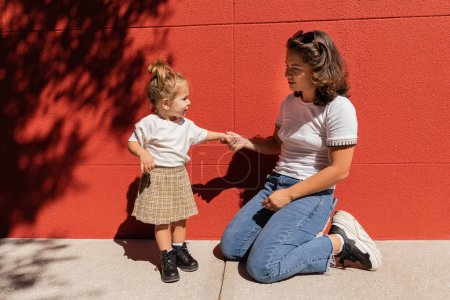 brünette Mutter in Jeans hält Händchen mit Kleinkind-Tochter, während sie in der Nähe der roten Wand sitzt 