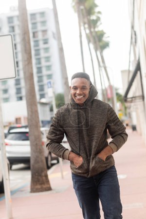 cheerful african american man in hoodie walking with hands in pockets in Miami 