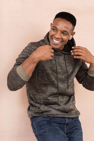fröhlicher afrikanisch-amerikanischer Mann in drahtlosem Kopfhörer, der nahe der Wand Musik hört 