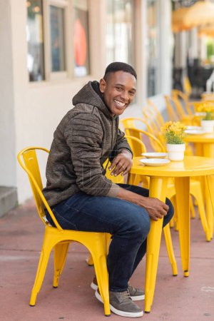 hombre afroamericano feliz en sudadera con capucha y jeans sentado en la mesa del bistro en la cafetería al aire libre en Miami 