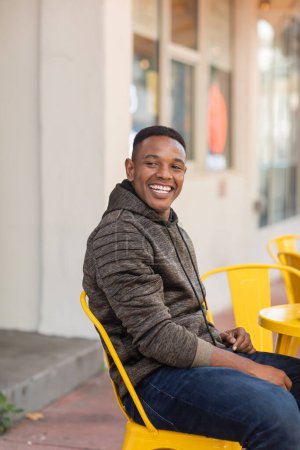 fröhlicher afrikanisch-amerikanischer Mann in Kapuzenpulli und Jeans sitzt am Bistrotisch im Outdoor-Café 