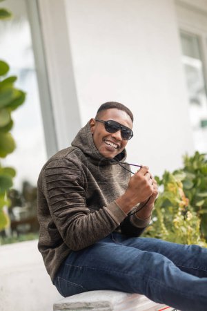 happy african american man in hoodie and sunglasses sitting on street in Miami 