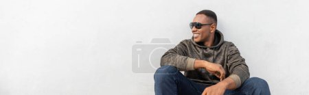 Téléchargez les photos : Heureux homme afro-américain dans des lunettes de soleil élégantes assis près du mur blanc, bannière - en image libre de droit