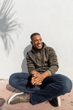 positiver afrikanisch-amerikanischer Mann in Jeans und Kapuzenpulli sitzt neben weißer Wand 
