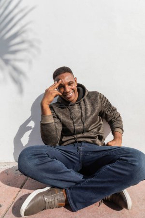 smiling african american man in jeans and hoodie sitting near white wall 