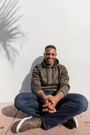 carefree african american man in jeans and hoodie sitting near white wall 