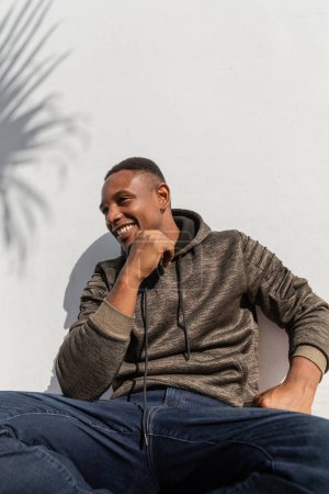 satisfied african american man in jeans and hoodie sitting near white wall 