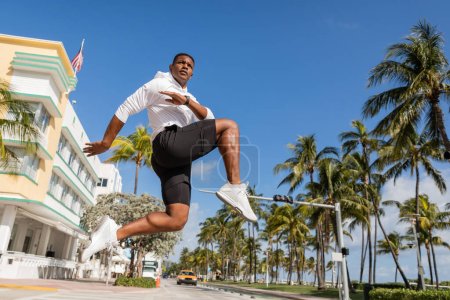 sportive african american man jumping next to palm trees and modern condominium in Miami 