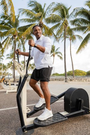 alegre afroamericano hombre ejercitando en cross trainer en al aire libre gimnasio 