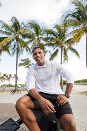 fröhlicher afrikanisch-amerikanischer Mann sitzt auf Crosstrainern in Outdoor-Fitnessstudio in Miami
