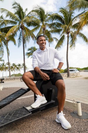 carefree african american sportsman sitting on cross trainer equipment in outdoor gym in Miami