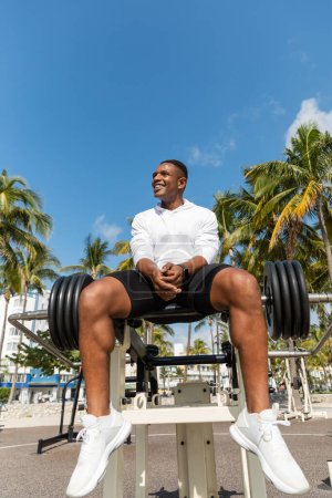 Blick auf einen fröhlichen afrikanisch-amerikanischen Sportler, der in der Nähe der Langhantel in einem Outdoor-Fitnessstudio in Miami sitzt 