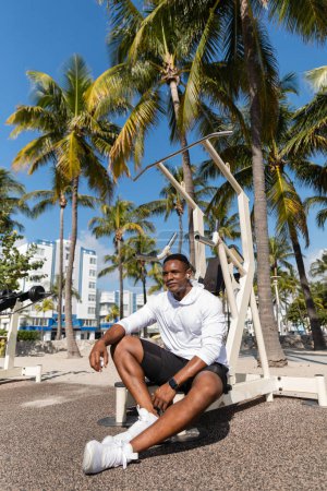 deportista afroamericano en auriculares inalámbricos descansando después del entrenamiento en Miami Beach 