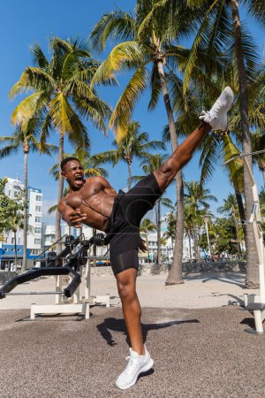 hombre afroamericano en pantalones cortos gritando mientras hace ejercicio de patada en Miami Beach 
