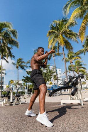 luchador afroamericano sin camisa en pantalones cortos haciendo ejercicio al lado de palmeras en Miami Beach 