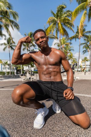 Téléchargez les photos : Sportif afro-américain fort en short et baskets montrant les muscles après l'entraînement à Miami plage - en image libre de droit