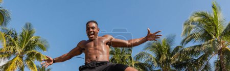 Foto de Vista de ángulo bajo del deportista afroamericano emocionado saltando cerca de palmeras verdes en Miami, pancarta - Imagen libre de derechos