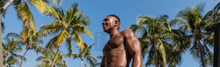 low angle view of cheerful african american sportsman standing next to palm trees in Miami, banner 