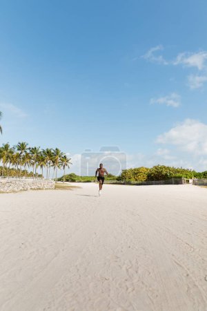 sportive and shirtless african american man in shorts running on sand in Miami beach