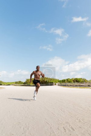 deportista afroamericano sin camisa en pantalones cortos que corren sobre arena en Miami Beach