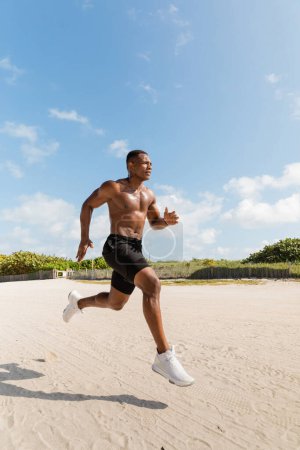 sportlicher und hemdsloser afrikanisch-amerikanischer Mann in kurzen Hosen beim Laufen auf Sand in Miami 