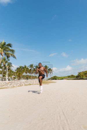 hombre afroamericano sin camisa en pantalones cortos corriendo sobre arena junto a palmeras verdes en la playa de Miami