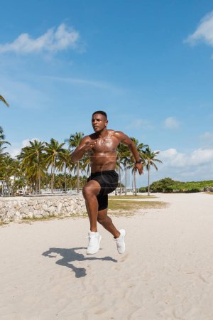 athletic african american man in shorts and sneakers running on sand next to palm trees in Miami beach