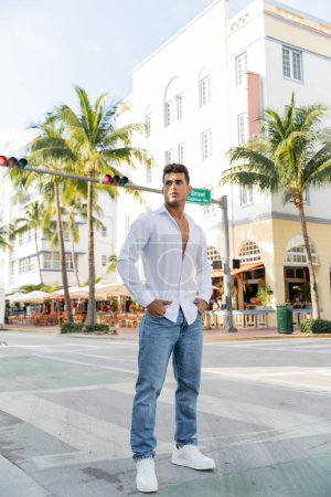 homme cubain en chemise blanche et jeans tenant la main dans les poches et debout sur la rue urbaine à Miami